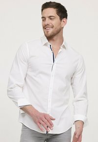 Chemise blanche à manches longues avec une patte de boutonnage intérieure bleu marine contrastante, un col boutonné et des poignets à un bouton, avec une texture subtile.