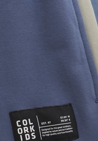 Bomulls tyg i nyanser av blått med en svart lapp som har vit text och koordinater. Slät struktur, avslappnad design och synliga sömnadsdetaljer.