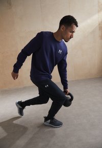 Mörkblå sweatshirt med en liten logotyp, svarta joggers; håller en svart kettlebell; grå träningsskor med överdel av mesh.