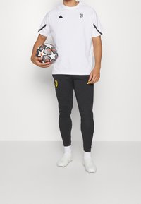 Biała koszulka sportowa z czarnymi akcentami, zestawiona z ciemnoszarymi spodniami o zwężanym kroju z żółtym logo. Zawodnik trzyma wzorzystą piłkę do nogi.