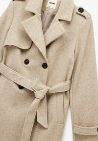 Beige wollen blend jas met dubbele zwarte knopen, grote kraag en een strikriem. Kenmerkt zich door een gestructureerd oppervlak en klepzakken.