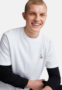 Mladý muž se usmívá, má na sobě bílé tričko s krátkým rukávem přes černou košili s dlouhým rukávem, sedí s rukama složenýma na klíně.