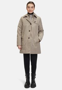 Beige trenchcoat med hög krage, stora knappar och två framfickor. Bärs över svarta byxor och kraftiga svarta stövlar. Slät textur.