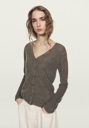 Cardigan marron à manches longues avec un col en V, doté de boutons sur le devant et d'une texture douce. Porté sur un pantalon ample de couleur claire.