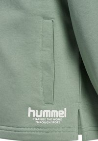 Groene sweatshirt met ribstructuur, voorzien van een zijvak en een wit "hummel" logo met daaronder "VERANDER DE WERELD DOOR SPORT".