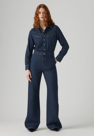 Combinaison en denim foncé avec un haut à col, poches à rabat et bas très large. Comprend des boutons sur le devant et une taille cintrée.