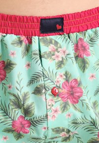 Shorts leggeri in tessuto verde menta con un motivo floreale composto da fiori rossi e rosa. Vita elastica con un'etichetta nera e un dettaglio di bottone rosso.