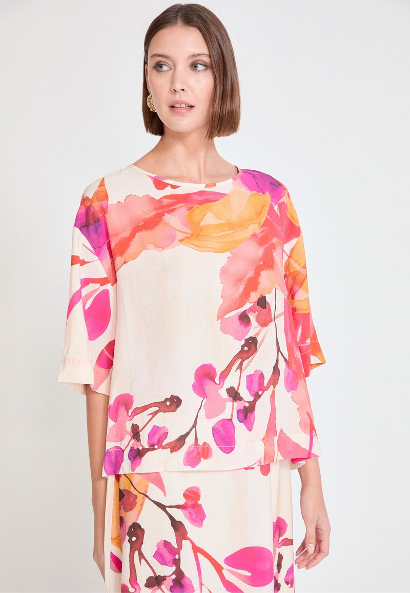 Blouse fleurie en tissu léger ; présente des motifs floraux vibrants rose et orange, des manches larges et un col rond.