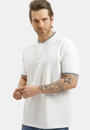 Mężczyzna z ułożonymi brązowymi włosami, ubrany w białą koszulkę polo z krótkim rękawem z prążkowanym kołnierzykiem i brzegami rękawów, pokazujący tatuaże na lewym przedramieniu.