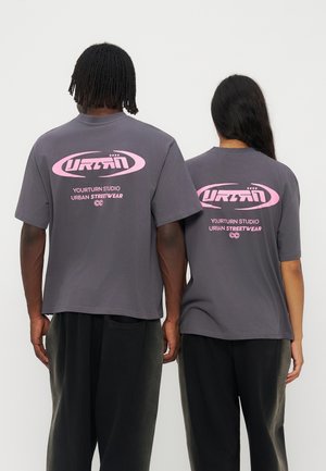 Deux personnes se tiennent côte à côte, dos tourné, portant des t-shirts gris avec du texte et un motif rose "URBAN Yourturn Studio Urban Streetwear" au dos.
