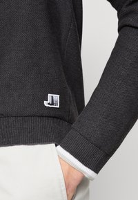 Ciemnoszary sweter z teksturowanej tkaniny, z białym wykończeniem mankietów i małą metką z logo na końcu, z widoczną ręką włożoną w kieszeń.