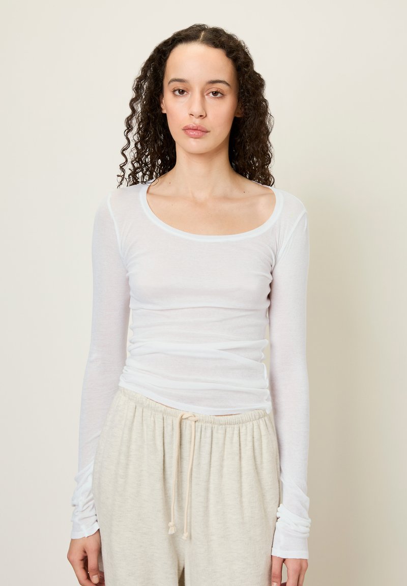 Top bianco a maniche lunghe aderente, realizzato in tessuto leggero, con scollatura a barca e dettagli arricciati in vita e sulle maniche.