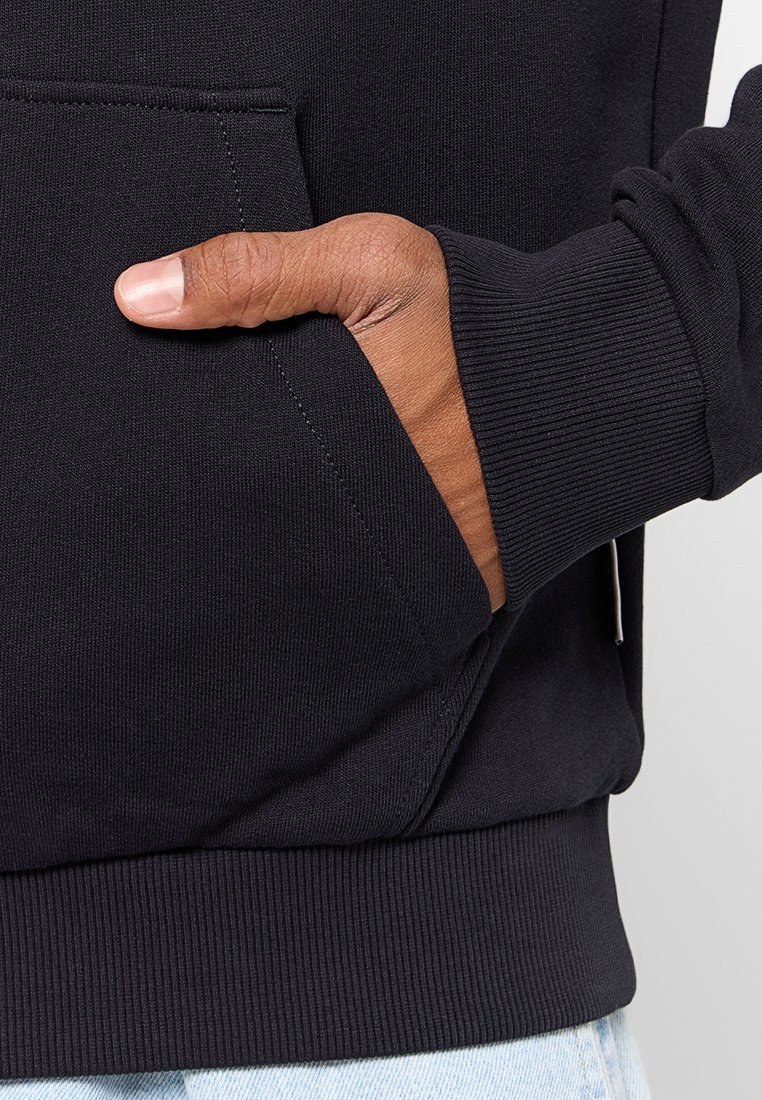 Prawa ręka umieszczona w przedniej kieszeni czarnego dresu z prążkowanymi mankietami, widoczna na tle jasnoniebieskich dżinsów.