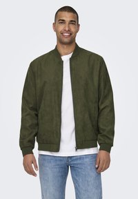 Giacca bomber in camoscio verde oliva con zip frontale, polsini e orlo a costine, indossata sopra una T-shirt bianca, abbinata a jeans azzurri.