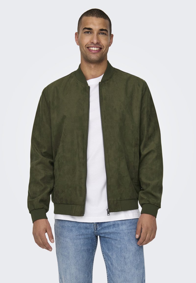 Giacca bomber in camoscio verde oliva con zip frontale, polsini e orlo a costine, indossata sopra una T-shirt bianca, abbinata a jeans azzurri.