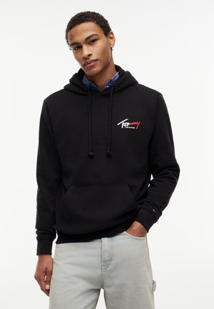 Νεαρός άνδρας με μαύρη κουκούλα με το λογότυπο "Tommy Jeans", μπλε καρό πουκάμισο από κάτω και ανοιχτό γκρι παντελόνι, στέκεται μπροστά σε λευκό φόντο.