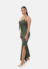 Mono verde oliva con un diseño ajustado, detalle de nudo en el frente, escote profundo y altas abertura laterales. Llevado con sandalias blancas de tacón alto.