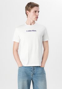 Balta medvilninė marškinė su trumpomis rankovėmis ir apvalia iškirpte. Krūtinės srityje yra purpurinė "Calvin Klein" logotipas. Dėvima su šviesiai mėlynomis džinsais.