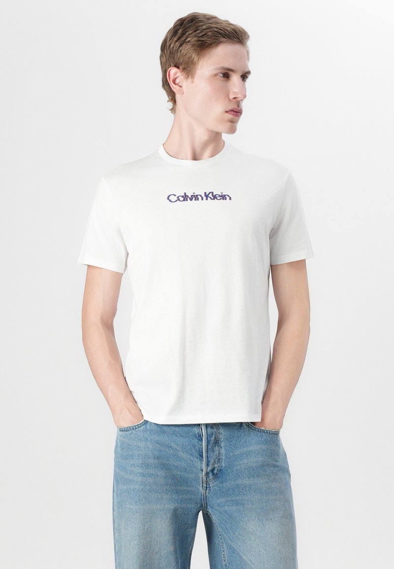 Camiseta de algodón blanca con mangas cortas y cuello redondo. Presenta un logo púrpura de "Calvin Klein" en el pecho. Se combina con jeans de un azul claro.