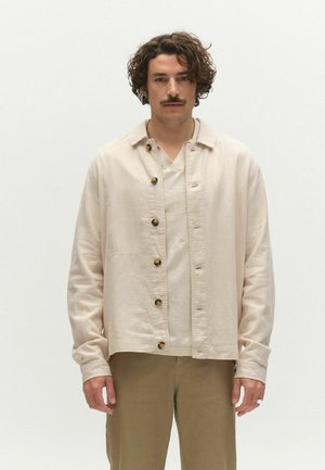 Kortsute ja vuntsidega mees, kes kannab beeži särki, millel on nööbid ja mis on seljas hele pluus ning pruunid püksid, seisab ühevärvilise valge tausta ees.