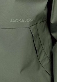 Giacca verde oliva con texture liscia, cuciture sottili e un logo JACK&JONES ben visibile sul davanti. Design minimalista.