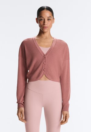 Žena nosící práškově růžový žebrovaný kardigan na knoflíky přes sladěný přiléhavý top a vysokým pasem legíny, stojící před jednobarevným pozadím.