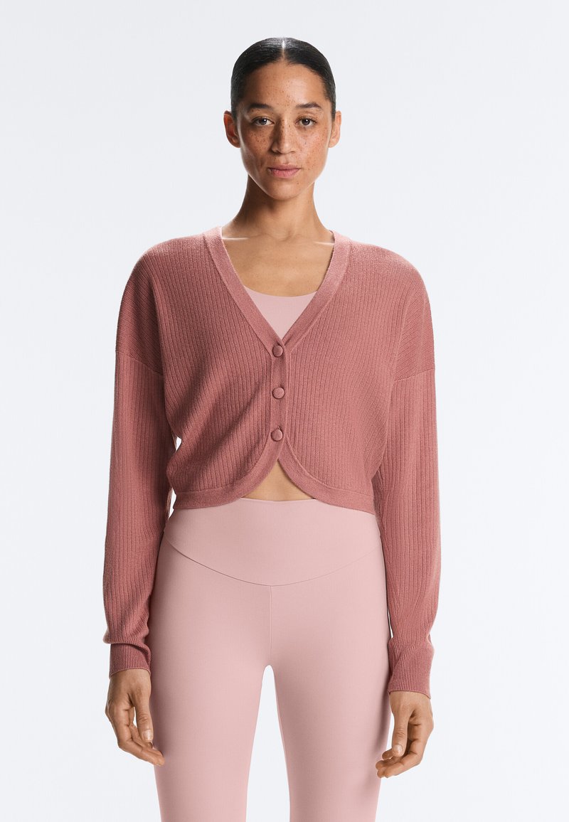 Femme portant un cardigan côtelé rose poussiéreux avec des boutons, sur un haut assorti ajusté et un legging taille haute, debout devant un fond uni.