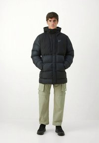 Chaqueta negra acolchada con cuello alto y capucha, con cierre frontal de cremallera y ligero brillo. Combinada con pantalones cargo oliva y zapatos negros.