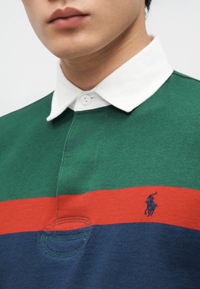 Άνδρας που φοράει ριγέ ράγκμπι πουκάμισο σε πράσινο, πορτοκαλί και ναυτικό μπλε χρώμα με λευκό γιακά και μικρό κεντημένο λογότυπο παίκτη πόλο στο στήθος.
