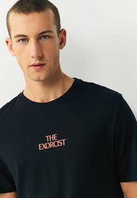 Melna īsām piedurknēm T-kreklu, kas izgatavots no kokvilnas, ar apaļu dekoltē. Uz kreisās krūtis ir "THE EXORCIST" logo korālī.