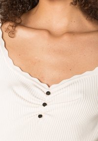 Top bianco a costine con scollatura smerlata, tre bottoni neri al centro e texture liscia con dettagli arricciati sotto i bottoni.