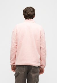 Άτομο με κοντά καστανά μαλλιά που φοράει ένα ανοιχτό ροζ φλις μπουφάν και σκούρο γκρι παντελόνι cargo, όρθιο με την πλάτη προς τα εμπρός μπροστά σε απλό φόντο.