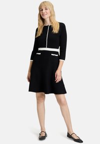 Robe noire avec un corsage ajusté, une ceinture blanche, des manches trois-quarts et deux poches avant, ornée de boutons noirs et d'une texture lisse.