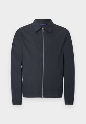 Mand stående med hænderne i lommerne iført en navyblå lynlåstrøje, blå skjorte, beige bukser og hvide sneakers på en hvid baggrund.