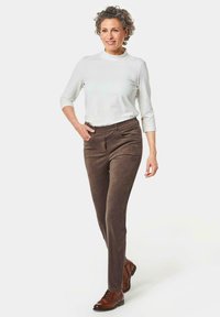 Bruine, suède-achtige slim fit broeken met twee voorzakken, gecombineerd met een witte blouse met lange mouwen. Model draagt bruine veterschoenen.