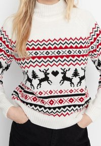 Weißer Strickpullover mit roten und schwarzen Zickzackmustern, Rentieren, Bäumen und Herzmotiven, getragen von einer Person mit Händen in den Taschen.