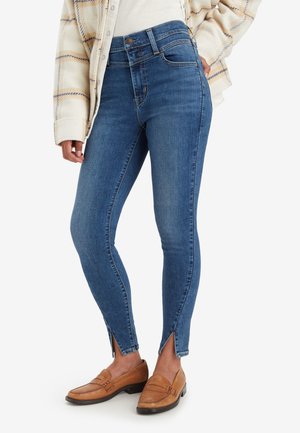 Frau trägt blaue Skinny-Jeans mit Reißverschluss-Schlitzen vorne, beige Mokassins und eine cremfarbene karierte Jacke über einem weißen Oberteil.