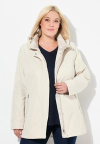 Blondýnka s béžovou bundou přes černý top, zapínající přední zip, na jednolitém bílém pozadí.