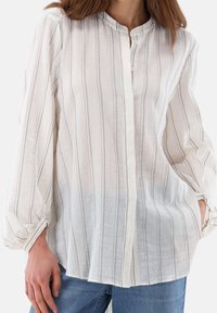 Chemise en lin blanc à fines rayures grises, encolure arrondie, fermeture par boutons, manches larges avec poignets froncés. Coupe décontractée avec texture légère.