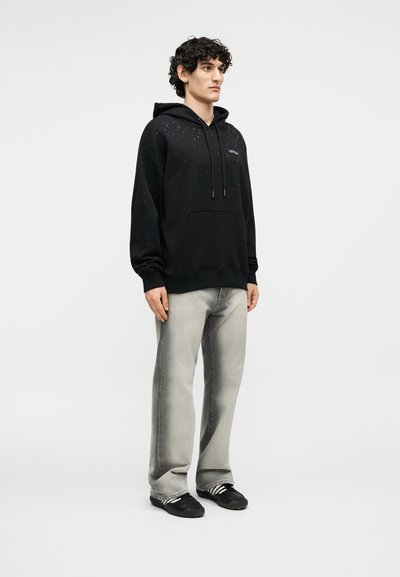 Jauns vīrietis stāv, nēsājot melnu kapuci ar baltām pērlītēm, gaiši pelēnas džinsas un melnas sporta kurpes ar baltām svītrām.