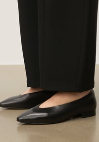 Ballerines en cuir noir avec un bout pointu et un détail découpé. Talon bloc bas. Portées avec un pantalon noir sur mesure. Texture lisse et design épuré.
