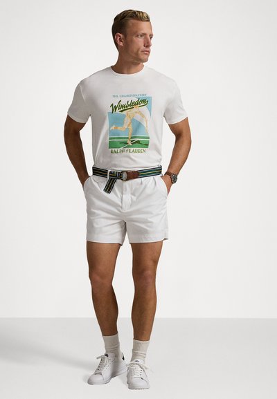 Weißes Baumwoll-T-Shirt mit Wimbledon-Grafik, weiße Shorts, gestreifter Gürtel und weiße Sneaker. Zubehör umfasst eine Uhr und helle Socken.