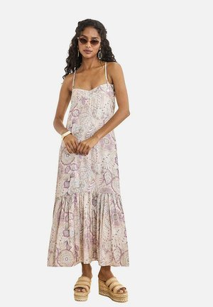 Mujer con cabello rizado, usando gafas de sol, un vestido maxi sin mangas con estampado floral, grandes pendientes de aro y sandalias con plataforma trenzadas, de pie con las manos entrelazadas.