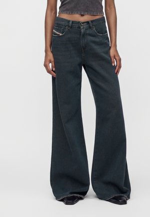 Persona che indossa jeans a gamba larga in denim scuro e un top corto grigio scuro, in piedi con le mani rilassate lungo i fianchi.