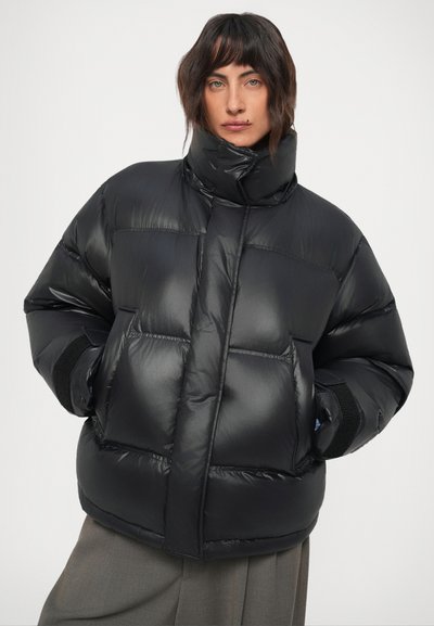 Osoba nosi crnu oversized puffer jaknu s visokim ovratnikom, ruke u džepovima, uparenu s labavim sivim hlačama na jednostavnoj pozadini.