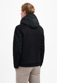 Zwarte, gestructureerde hoodie met een gevoerde zwarte capuchon, voorzien van een subtiel logo op de linker schouder en ribgebreide manchetten, gecombineerd met lichtgekleurde broek.