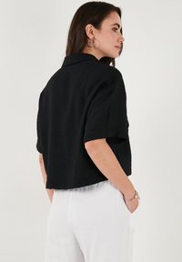 Maglietta nera corta con una vestibilità rilassata, maniche corte e colletto. Indossata con pantaloni bianchi strutturati, con una finitura in tessuto liscio.