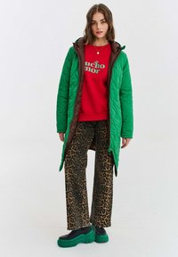 Zelený prešívaný dlhý kabát s hnedými akcentami, červená mikina s textom, široké nohavice s leopardím vzorom a masívne zelené platformové topánky.