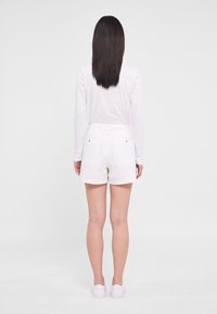 Short en coton blanc avec deux poches arrière et boutons dorés, vu de dos. Associé à un haut blanc à manches longues et des chaussures blanches.