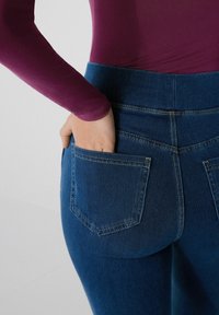 Jeans de mezclilla azul con alta cintura, que cuentan con dos bolsillos traseros y costuras amarillas en contraste. Combinados con una blusa morada de manga larga.